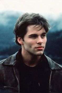 a young man in a leather jacket looking at the camera with mountains in the background