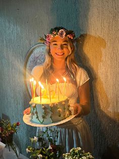 a woman holding a cake with lit candles on it in front of flowers and greenery