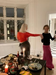 two women dancing around a table full of food and drinks