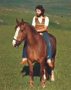 a man riding on the back of a brown horse in a green field with mountains behind him