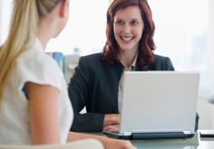 a woman sitting at a desk with a laptop computer in front of her, talking to another woman