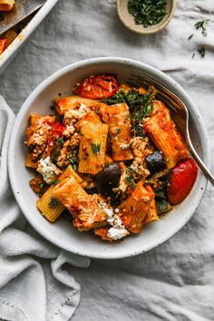 a bowl filled with pasta and vegetables on top of a white cloth next to a spoon