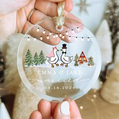 a person holding up a glass ornament with two geese on it and christmas lights in the background