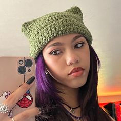 a woman with purple hair holding up a cell phone wearing a green crochet hat