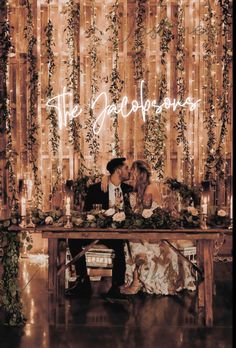 a bride and groom sitting at a table in front of a curtain with lights on it