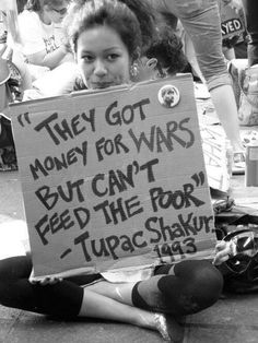 a woman sitting on the ground holding a sign that says they got money for wars but can't feed the poor tupac shaka