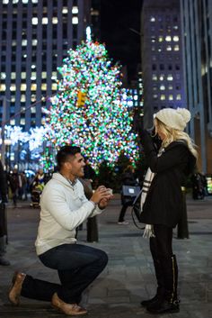 a man kneeling down next to a woman in front of a lit up christmas tree