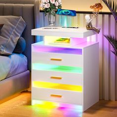 a white night stand with multicolored lights on it next to a bed and flowers