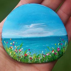 a painted rock in the palm of someone's hand with flowers and grass on it