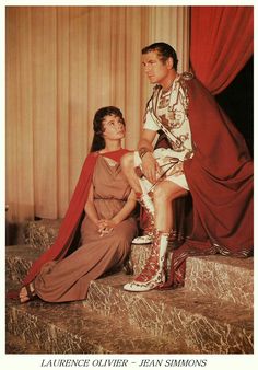 a man and woman sitting on top of a bed next to each other in front of red curtains