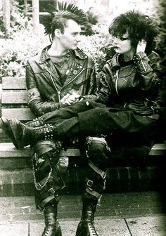 two people are sitting on a bench wearing punk clothes and leather jackets, one is holding his leg up to the other's chest