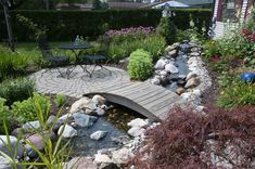 a wooden bridge over a small stream in a garden area with flowers and plants around it