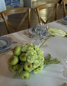 an arrangement of green apples and flowers on a white table cloth with silverware in the background