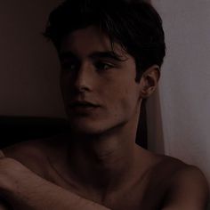 a shirtless young man sitting on a bed with his arm around the head and looking off into the distance