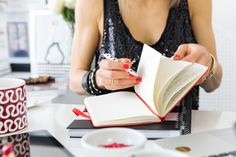 a woman sitting at a table with an open book in front of her and holding a pen