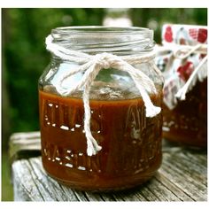 two jars filled with homemade barbecue sauce on top of a wooden table in front of some trees