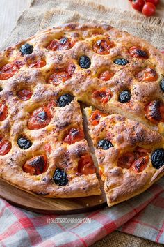 a pizza with olives and tomatoes cut in half on a wooden cutting board next to a red checkered napkin