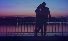 two people standing next to each other in front of a body of water at sunset