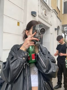 a woman taking a photo with her cell phone while wearing a leather jacket and holding a green bottle