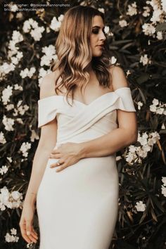 a woman standing in front of flowers wearing a white dress with off the shoulder sleeves