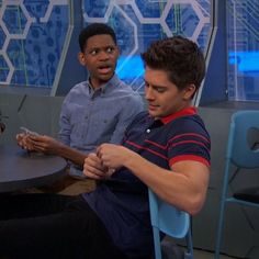 two young men sitting at a table in front of blue chairs, one holding a cell phone