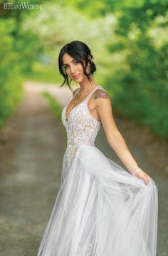 a beautiful woman in a white dress posing for a photo on a dirt road with trees behind her