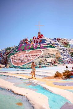 a woman walking across a sandy beach next to a hill covered in colorfully painted hills