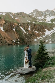 a bride and groom standing on top of a rock next to a lake