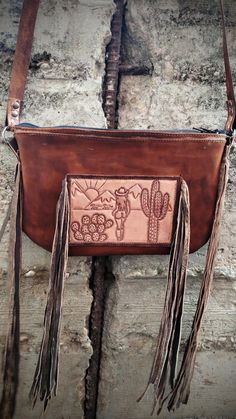 Tooled leather crossbody purse featuring a beautiful desert scene with a lonely cowgirl walking through. This purse has fringe on either side of the purse and on the front with the tooled leather piece.  Hand Tooled leather strap in a floral design. Western Style Crossbody Shoulder Bag For Travel, Western Style Hand Tooled Shoulder Bag For Travel, Western Leather Crossbody Shoulder Bag, Leather Hand Tooled Shoulder Bag For Western-themed Events, Western Style Crossbody Bags For Western-themed Events, Western Leather Bags With Concho Details, Bohemian Brown Bags With Concho, Bohemian Leather Bag With Concho, Beautiful Desert