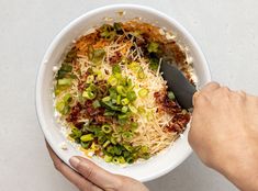 a person holding a bowl of food with shredded cheese and green onions in it, while another hand holds a spoon over the bowl