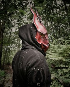 a man wearing a red mask and black jacket in the woods with trees behind him