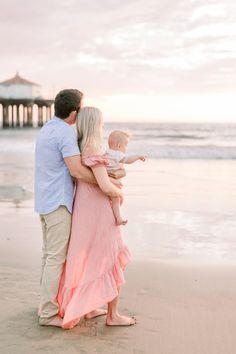 a man and woman holding a baby on the beach at sunset with pier in background