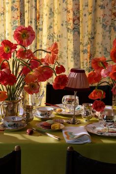 the table is set with tea cups, plates and vases filled with red flowers