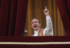 a man standing at a podium with his hand in the air and pointing to something