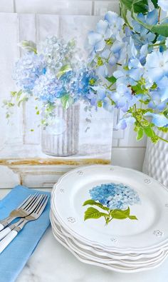 blue hydrangeas are in a vase next to white plates and silverware