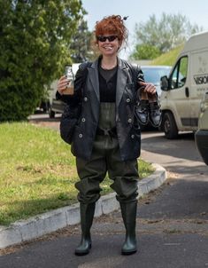 a woman with red hair wearing green pants and black jacket holding a drink in her hand