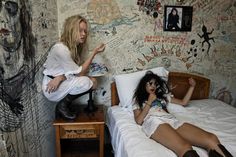 two women sitting on top of a bed in a room with walls covered in graffiti