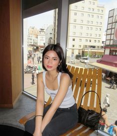 a young woman sitting on top of a wooden bench next to a large glass window