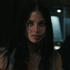 a woman with long hair and dark makeup looks at the camera in a dimly lit room