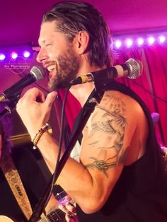 a man singing into a microphone while holding a guitar in his right hand and wearing a black vest