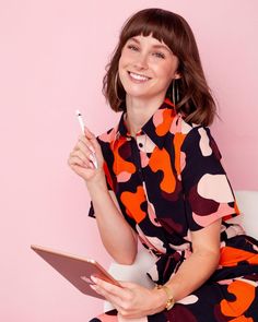 a woman sitting on a white chair holding a tablet and smiling at the camera while wearing a colorful dress