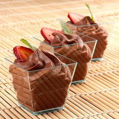 three desserts with chocolate frosting and strawberries in small cups on a bamboo mat