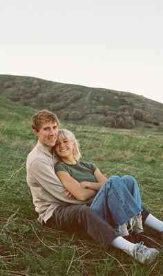 a man and woman sitting on the ground in front of a green hill with trees