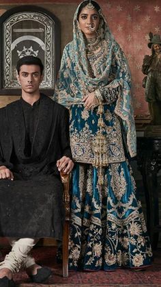 a man and woman sitting on a chair in front of a wall with ornate paintings