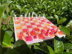 a checkerboard board is sitting in the middle of green plants and leaves, with small plastic chess pieces on it