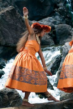 two women in orange dresses are walking on rocks near the water and some rocks, one woman is holding her hand up