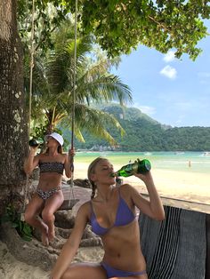 two women in bikinis sitting on the beach drinking from green bottles and talking to each other