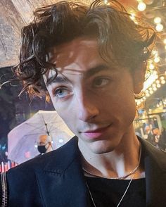 a close up of a person wearing a suit and necklace with an umbrella in the background