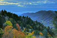 the mountains are covered in colorful trees and surrounded by blue sky with white clouds above them