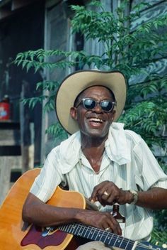 a man wearing sunglasses and a hat playing an acoustic guitar in front of a tree